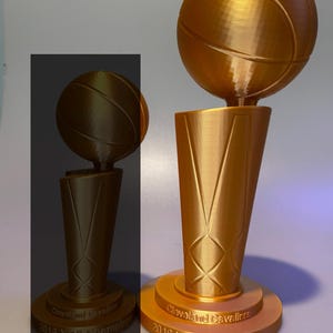 May include: A gold-coloured 33 cm Cleveland Cavaliers 2016 NBA Champions trophy featuring a basketball mounted on a tapered base. The base is circular with the text Cleveland Cavaliers 2016 NBA Champions embossed on the front surface.