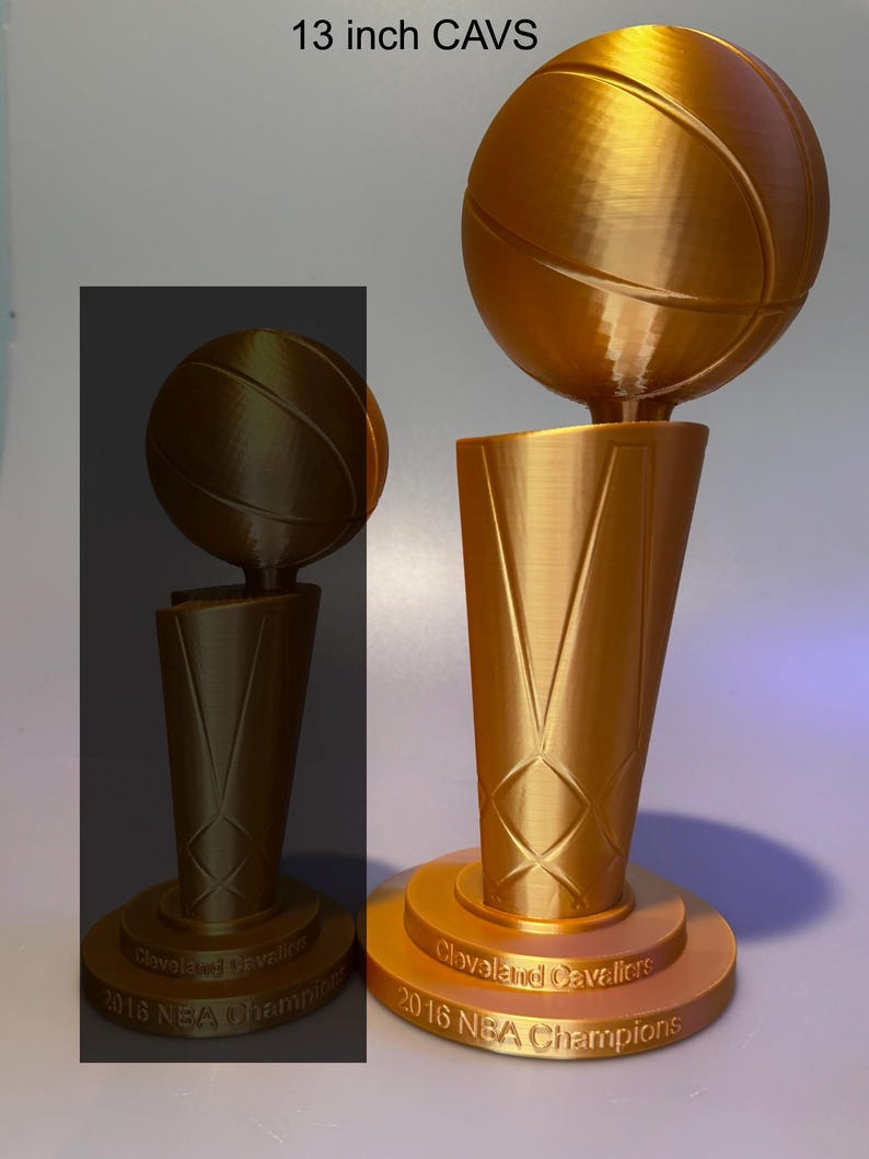 May include: A gold-coloured 33 cm Cleveland Cavaliers 2016 NBA Champions trophy featuring a basketball mounted on a tapered base. The base is circular with the text Cleveland Cavaliers 2016 NBA Champions embossed on the front surface.