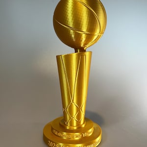 May include: A gold-coloured trophy featuring a basketball mounted on a tapered, net-patterned base. The circular pedestal base is embossed with the text Oklahoma City Thunder 2025 NBA Champions in a raised, metallic gold font against the surface.
