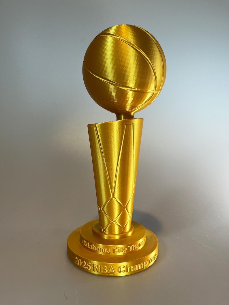 May include: A gold-coloured trophy featuring a basketball mounted on a tapered, net-patterned base. The circular pedestal base is embossed with the text Oklahoma City Thunder 2025 NBA Champions in a raised, metallic gold font against the surface.