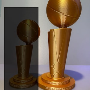 May include: A gold-coloured 33 cm Oklahoma City Thunder 2025 NBA Champions trophy featuring a basketball mounted on a tapered base. The base is engraved with the text Oklahoma City Thunder 2025 NBA Champions. Two versions are shown.