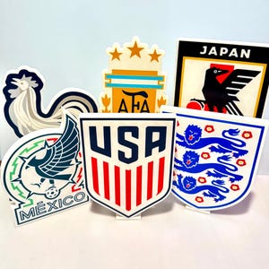 May include: A collection of colorful, stylized logos representing various national soccer teams. The logos include the USA, Mexico, Japan, England, Argentina, and France. Each logo is a unique shape and design, with vibrant colors and distinct imagery.