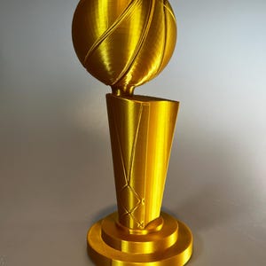 May include: A gold-coloured trophy featuring a basketball perched atop a tapered, cylindrical base. The object has a smooth, metallic finish with visible 3D-printed layer lines throughout the structure, including the round pedestal base.