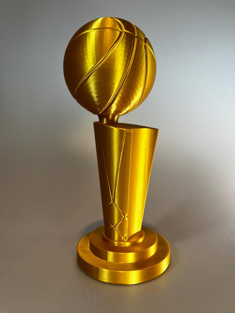 May include: A gold-coloured trophy featuring a basketball perched atop a tapered, cylindrical base. The object has a smooth, metallic finish with visible 3D-printed layer lines throughout the structure, including the round pedestal base.
