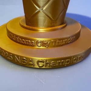 May include: A gold-coloured trophy base with a tiered circular design. The upper tier features the text Oklahoma City Thunder, while the lower tier displays the text 2025 NBA Champions. The surface has a visible 3D printed texture throughout.