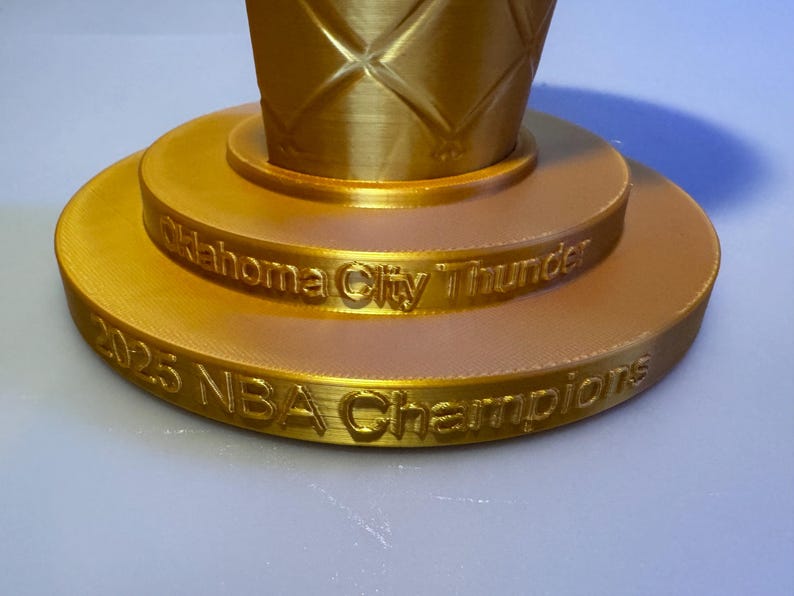 May include: A gold-coloured trophy base with a tiered circular design. The upper tier features the text Oklahoma City Thunder, while the lower tier displays the text 2025 NBA Champions. The surface has a visible 3D printed texture throughout.