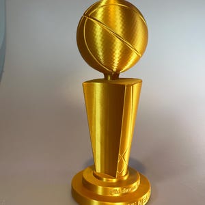 May include: A gold-coloured trophy featuring a basketball mounted on a tapered, multi-faceted pedestal base. The base includes the embossed text Oklahoma and 2025 NEA. The entire object has a metallic finish with visible 3D printed layer lines.