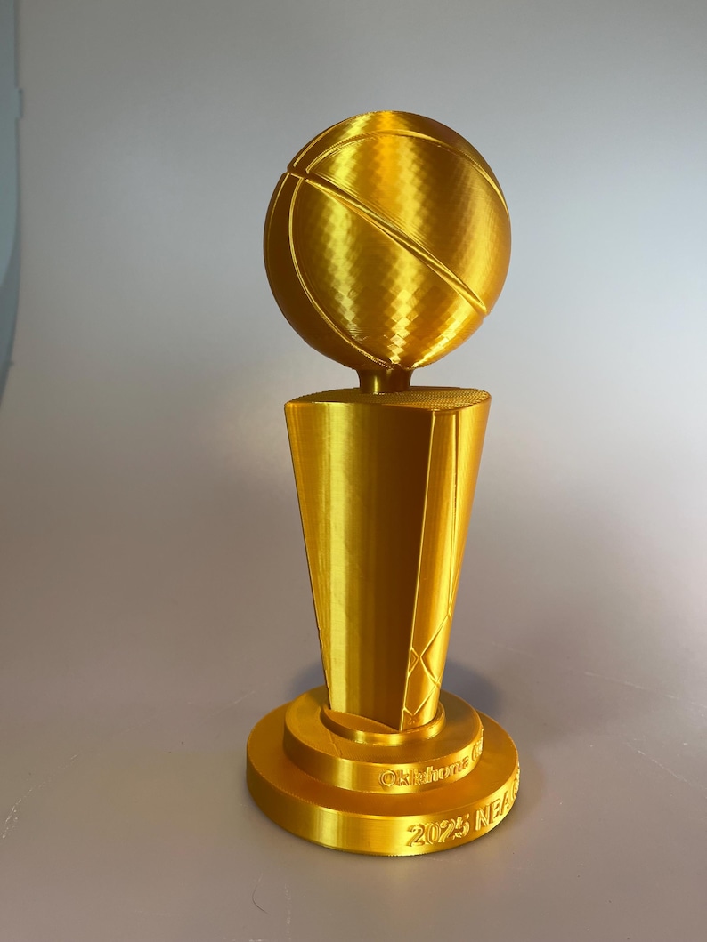 May include: A gold-coloured trophy featuring a basketball mounted on a tapered, multi-faceted pedestal base. The base includes the embossed text Oklahoma and 2025 NEA. The entire object has a metallic finish with visible 3D printed layer lines.