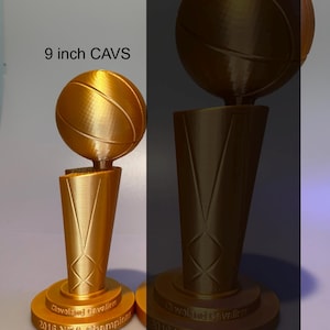May include: Two gold-coloured 3D printed trophies featuring a basketball atop a tapered base. The base is inscribed with "Cleveland Cavaliers 2016 NBA Champions". The smaller trophy is labelled "9 inch CAVS" in black text above it.
