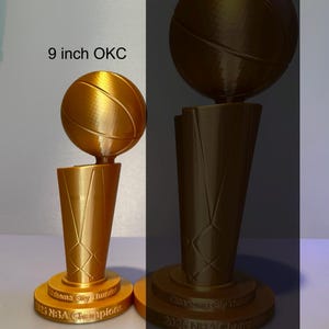 May include: Two gold-coloured 3D-printed basketball trophies featuring a basketball atop a tapered base. The base is inscribed with "Oklahoma City Thunder 2025 NBA Champions". One trophy is labelled as 9 inches tall, while the other is larger.