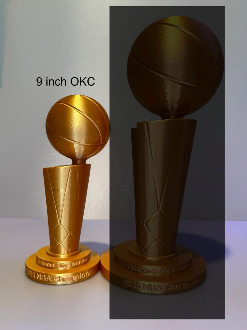 May include: Two gold-coloured 3D-printed basketball trophies featuring a basketball atop a tapered base. The base is inscribed with "Oklahoma City Thunder 2025 NBA Champions". One trophy is labelled as 9 inches tall, while the other is larger.