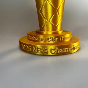 May include: A gold-coloured trophy with a diamond-patterned texture on the upper section. The base features two tiers with engraved text reading Oklahoma City Thunder on the top tier and 2025 NBA Champions on the bottom tier.