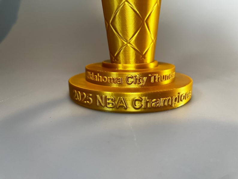 May include: A gold-coloured trophy with a diamond-patterned texture on the upper section. The base features two tiers with engraved text reading Oklahoma City Thunder on the top tier and 2025 NBA Champions on the bottom tier.