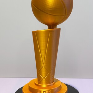 May include: A gold-coloured trophy featuring a basketball mounted on a tapered, textured base. The base includes the embossed text #1 Dad and Happy Father's Day 2026. The trophy has a smooth, metallic finish and stands on a circular black platform.