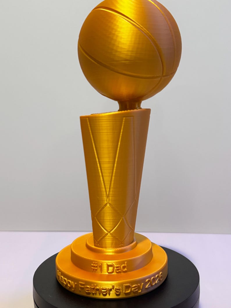 May include: A gold-coloured trophy featuring a basketball mounted on a tapered, textured base. The base includes the embossed text #1 Dad and Happy Father's Day 2026. The trophy has a smooth, metallic finish and stands on a circular black platform.