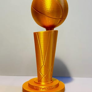May include: A gold-coloured trophy featuring a basketball mounted on a tapered, textured base. The base includes the engraved text New York Knicks and 2026 NBA Champions. The object has a visible 3D printed layered finish throughout its structure.