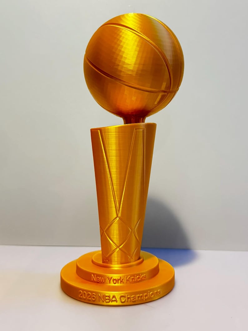May include: A gold-coloured trophy featuring a basketball mounted on a tapered, textured base. The base includes the engraved text New York Knicks and 2026 NBA Champions. The object has a visible 3D printed layered finish throughout its structure.