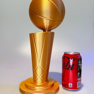 May include: A gold-coloured 3D-printed trophy featuring a basketball atop a tapered base. The base is inscribed with "Oklahoma City Thunder 2025 NBA Champions". A standard red soda can is positioned next to the 33 cm tall trophy for scale.