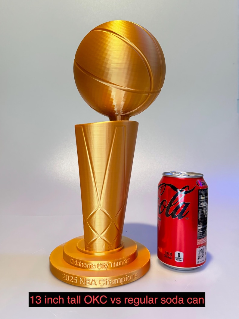 May include: A gold-coloured 3D-printed trophy featuring a basketball atop a tapered base. The base is inscribed with "Oklahoma City Thunder 2025 NBA Champions". A standard red soda can is positioned next to the 33 cm tall trophy for scale.