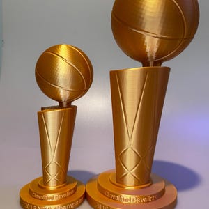 May include: Two gold-coloured trophies featuring a basketball atop a tapered base. Each base is inscribed with the text Cleveland Cavaliers 2016 NBA Champions. The trophies are identical in design but differ in size, with one being noticeably larger.