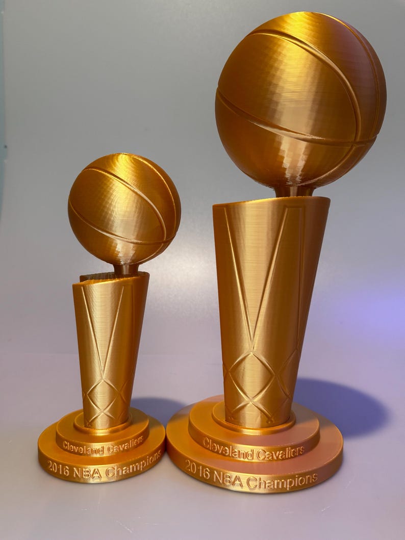 May include: Two gold-coloured trophies featuring a basketball atop a tapered base. Each base is inscribed with the text Cleveland Cavaliers 2016 NBA Champions. The trophies are identical in design but differ in size, with one being noticeably larger.