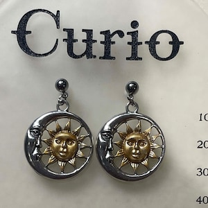 May include: A pair of silver-tone earrings featuring a sun and moon design. The sun is gold-colored and centered within a silver-tone circle, with a crescent moon on the left side. The word "Curio" is printed at the top.