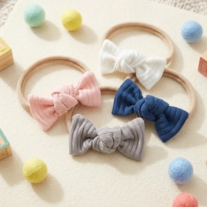 May include: Five baby headbands with ribbed bow details in white, pink, grey, and navy blue. Each bow is attached to a neutral elastic band. Scattered around are colourful felt balls in yellow, blue, and pink, creating a playful arrangement.
