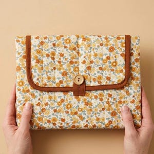 May include: A floral-patterned clutch with a brown trim and button closure. The clutch features a quilted design with a repeating pattern of yellow and orange flowers on a cream background. The trim and button loop are brown.