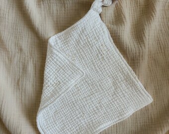 Peut inclure: Une couverture en mousseline de coton blanche texturée pour bébé avec une sucette attachée. La couverture est posée sur une surface texturée beige.