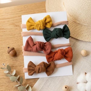 May include: Five elastic headbands featuring knotted fabric bows in mustard yellow, dark green, terracotta, burnt orange, and brown. The headbands are displayed on a white rectangular card, showcasing a ribbed texture on each bow.