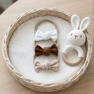 May include: Three baby headbands with knotted bows in white, brown, and beige, presented on a white linen surface. A white crochet bunny rattle with wooden ring sits beside the headbands, all within a woven basket.