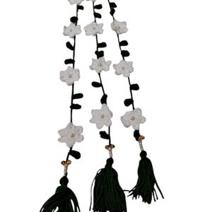 May include: A decorative hanging ornament featuring three vertical strands of dark green yarn adorned with white crocheted flowers and small central beads. Each strand terminates in a dark green tassel with gold and white bead accents. The letter D is visible.