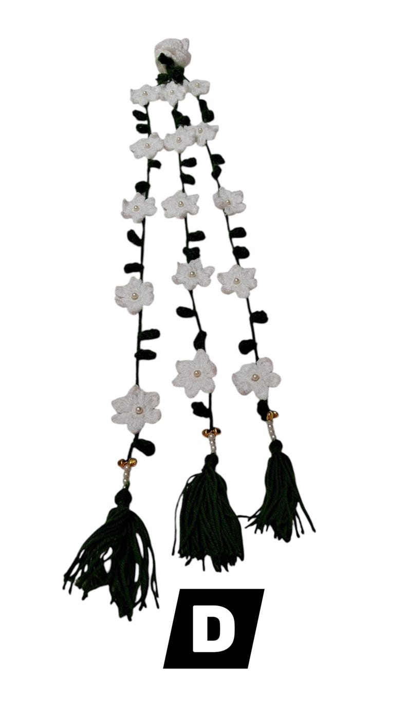 May include: A decorative hanging ornament featuring three vertical strands of dark green yarn adorned with white crocheted flowers and small central beads. Each strand terminates in a dark green tassel with gold and white bead accents. The letter D is visible.