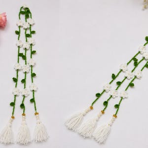May include: A long, vertical crochet garland featuring three green yarn vines adorned with small white flowers and green leaves. Each vine terminates in a gold-toned bead, a small white pearl-like bead, and a thick white yarn tassel at the base.