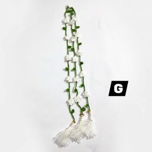 May include: A long, crocheted garland featuring a thin green vine with small white five-petaled flowers attached at intervals. The garland ends in three white thread tassels with small gold-toned beads at the top. The letter G is visible.