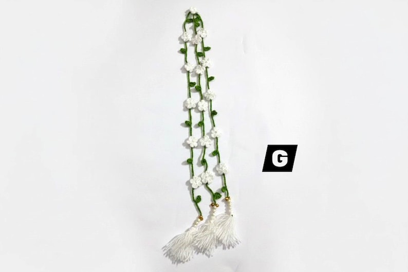 May include: A long, crocheted garland featuring a thin green vine with small white five-petaled flowers attached at intervals. The garland ends in three white thread tassels with small gold-toned beads at the top. The letter G is visible.