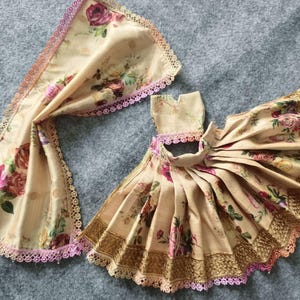 Puede incluir: Un conjunto de vestido y bufanda para muñeca beige. La tela presenta un estampado floral con rosas rosas y burdeos. El vestido tiene una falda plisada y ribete dorado. Ambas piezas están ribeteadas con encaje rosa.