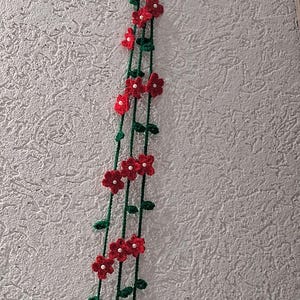 May include: A hanging crochet decoration featuring three long green yarn stems adorned with small red flowers. Each flower has five petals and a tiny white bead at its center. The stems include small green leaves and are attached to a light green hook.