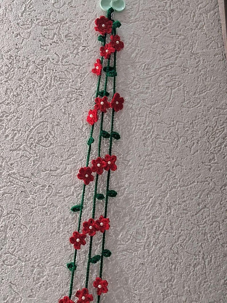 May include: A hanging crochet decoration featuring three long green yarn stems adorned with small red flowers. Each flower has five petals and a tiny white bead at its center. The stems include small green leaves and are attached to a light green hook.