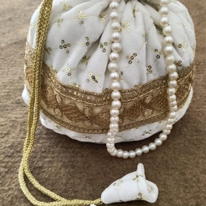 Może przedstawiać: Biała torebka ze sznurkiem do ściągania, haftem złotym i naszyjnikiem z pereł. Torebka ma aksamitną fakturę i jest ozdobiona złotymi cekinami.