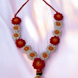 Könnte beinhalten: Eine handgefertigte Blumengirlande mit roten, weißen und orangefarbenen gehäkelten Blumen. Die Girlande hat eine rote Kordel und eine dekorative Quaste mit orangefarbenen Perlen. Die Blumen haben eine perlenartige Mitte.