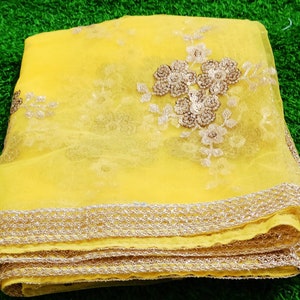 May include: A folded, sheer yellow fabric with intricate gold floral embroidery and a delicate gold trim along the edge. The fabric is layered, showcasing the detailed design and texture. The background is a vibrant green.