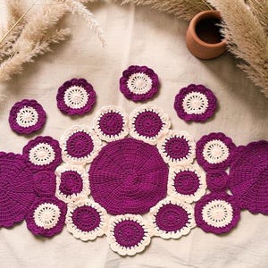 Peut inclure: Ensemble d'articles crochetés à la main, violets et crème. L'ensemble comprend une grande pièce maîtresse circulaire, des dessous de verre et des sets de table, tous avec un motif floral. Les articles sont disposés sur une surface neutre, avec des plantes séchées et un petit pot en terre cuite en arrière-plan.