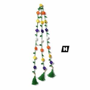 May include: A hanging decoration featuring three long green crocheted vines adorned with small yellow, orange, purple, and white flowers. Each vine terminates in a green yarn tassel and a small bead. The letter H is visible in a black square.