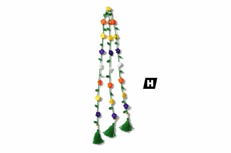 May include: A hanging decoration featuring three long green crocheted vines adorned with small yellow, orange, purple, and white flowers. Each vine terminates in a green yarn tassel and a small bead. The letter H is visible in a black square.