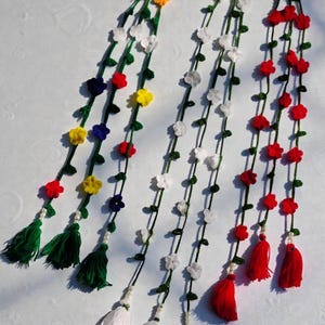 May include: Three decorative hanging garlands featuring crocheted green vines with small colorful flowers. The first has yellow, red, and blue flowers with green tassels. The second has white flowers with white tassels. The third has red flowers with red tassels.