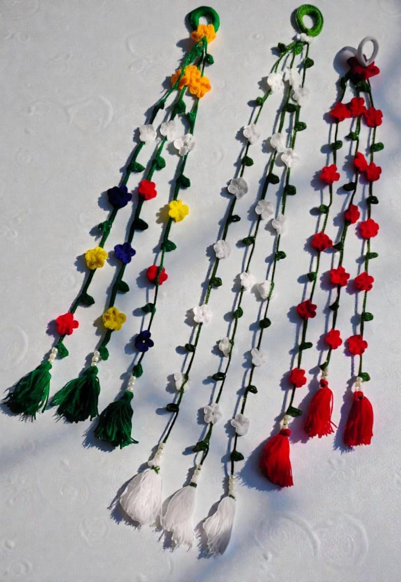 May include: Three decorative hanging garlands featuring crocheted green vines with small colorful flowers. The first has yellow, red, and blue flowers with green tassels. The second has white flowers with white tassels. The third has red flowers with red tassels.