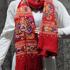 Peut inclure: Une écharpe rouge vif avec des broderies complexes dans diverses couleurs, dont le jaune, le violet et le vert. L'écharpe présente des motifs et des dessins détaillés, créant un textile riche visuellement. L'écharpe est drapée sur un vêtement blanc.