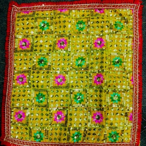 May include: A yellow fabric square with a repeating pattern of pink, green, and gold embroidered flowers. The fabric is decorated with sequins and has a red and gold border.