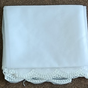 Op de afbeelding: Een gevouwen, doorschijnende witte stof met een delicate, geschulpte kanten rand langs één rand. De stof lijkt licht van gewicht en kan worden gebruikt voor bruidskleding of decoratieve doeleinden. De kanten rand voegt een vleugje elegantie toe.